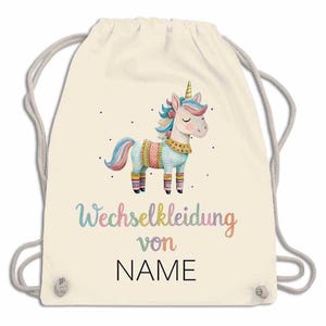 May include: A cream-colored drawstring backpack featuring a colorful unicorn illustration. The unicorn has a golden horn, a pink mane, and is wearing a patterned outfit. The text "NAME" is printed below the unicorn in black, with the text "Wechselkleidung von" above it.