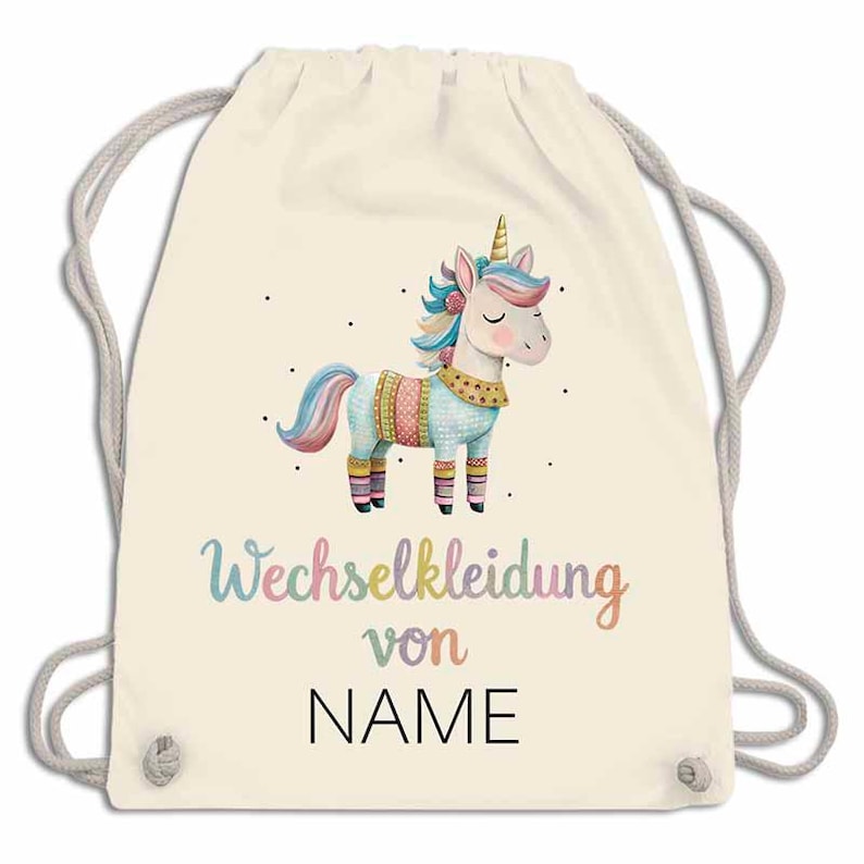 May include: A cream-colored drawstring backpack featuring a colorful unicorn illustration. The unicorn has a golden horn, a pink mane, and is wearing a patterned outfit. The text "NAME" is printed below the unicorn in black, with the text "Wechselkleidung von" above it.