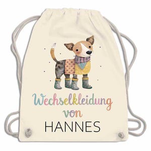 May include: Cream-colored drawstring backpack featuring a watercolor illustration of a dog wearing a sweater, scarf, and boots. The text "Wechselkleidung von HANNES" is printed below the dog in colorful script.