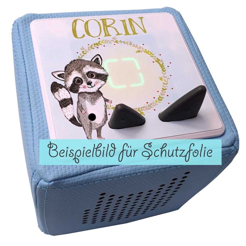 May include: A light blue cube-shaped speaker with a raccoon illustration and the name "CORIN" on the front. The speaker has two black buttons and a small digital display. The text "Beispielbild f&uuml;r Schutzfolie" is visible on the bottom of the speaker.