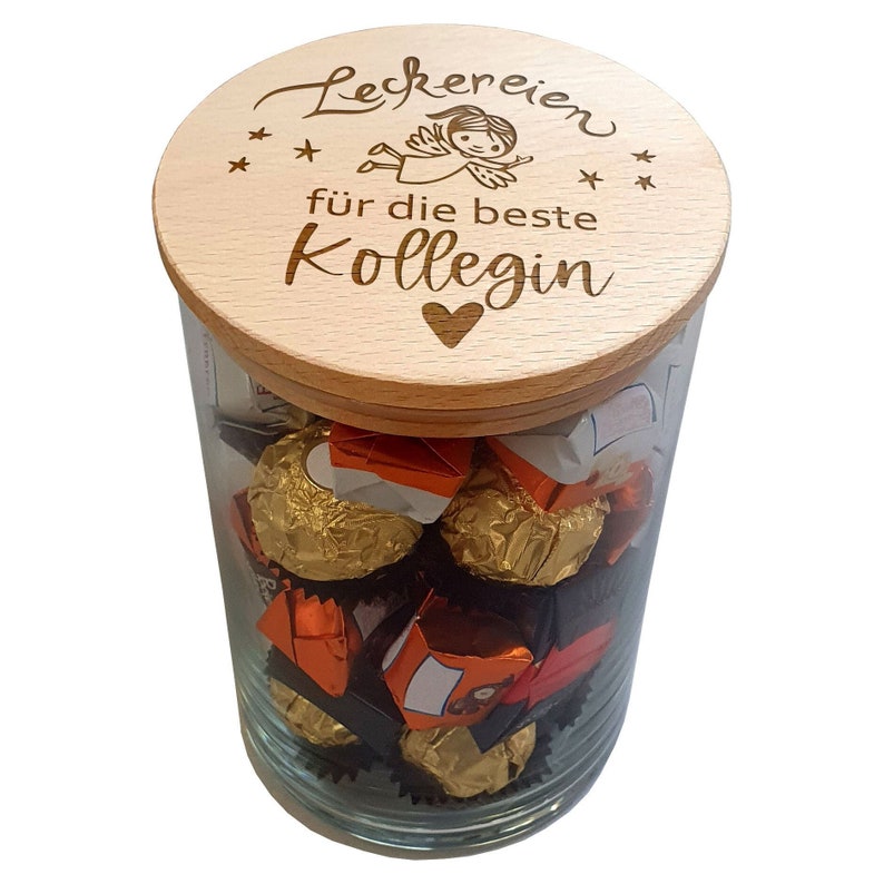May include: A clear glass jar filled with chocolates, topped with a wooden lid. The lid is engraved with the words "Leckereien f&uuml;r die beste Kollegin", a small angel, stars, and a heart. The jar contains various wrapped chocolates.