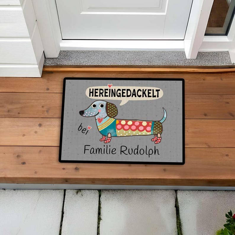 May include: Rectangular doormat with a cartoon dachshund design. The dog is multi-colored with a speech bubble above its head that says "HEREINGEDACKELT". The text "bei Familie Rudolph" is below the dog.