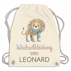 May include: A cream-colored drawstring backpack featuring a watercolor illustration of a lion wearing a striped scarf and socks. The text "Wechselkleidung von LEONARD" is written in colorful script below the lion.