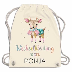 May include: A cream-colored drawstring backpack featuring a watercolor illustration of a deer wearing a scarf and sweater. The text "Wechselkleidung von RONJA" is written in colorful script below the deer.