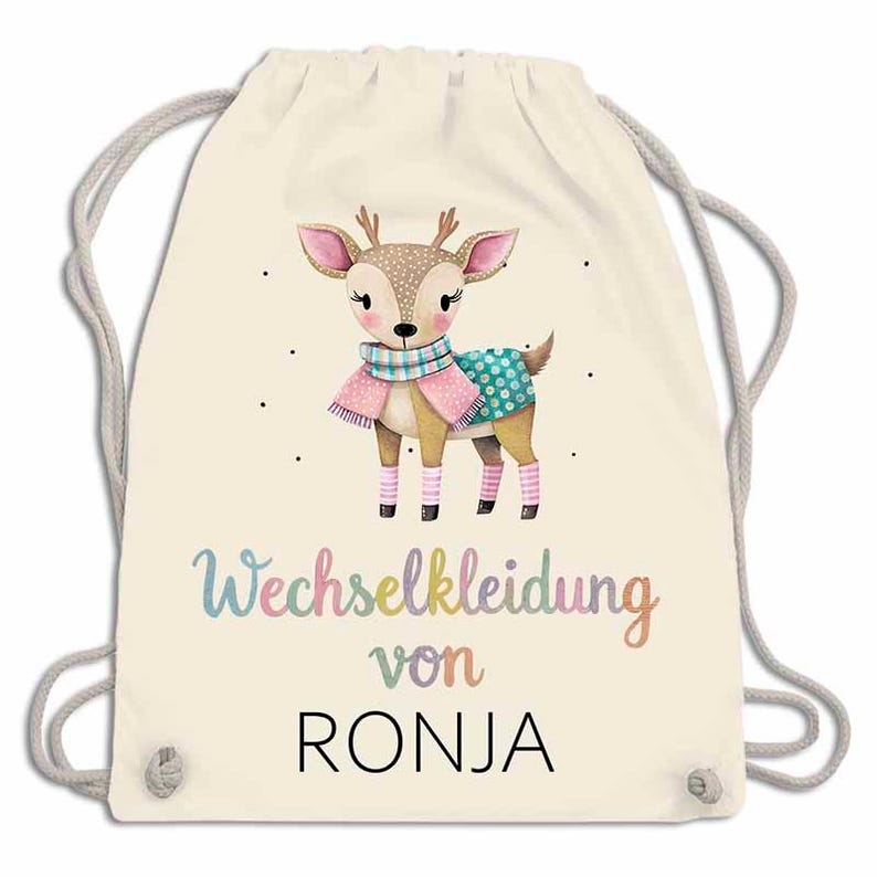May include: A cream-colored drawstring backpack featuring a watercolor illustration of a deer wearing a scarf and sweater. The text "Wechselkleidung von RONJA" is written in colorful script below the deer.