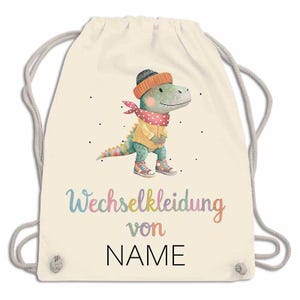 May include: A cream-colored drawstring backpack featuring a watercolor illustration of a dinosaur wearing a hat, scarf, and sweater. The text "Wechselkleidung von NAME" is written in colorful script below the dinosaur.