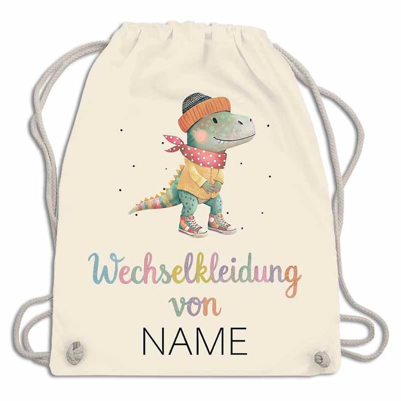 May include: A cream-colored drawstring backpack featuring a watercolor illustration of a dinosaur wearing a hat, scarf, and sweater. The text "Wechselkleidung von NAME" is written in colorful script below the dinosaur.