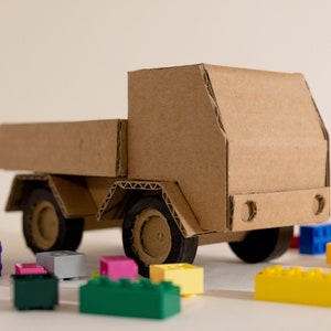 Può includere: Un camion giocattolo fatto a mano in cartone, con esterno marrone e ruote nere. Il camion è circondato da colorati mattoncini giocattolo di varie forme e dimensioni, suggerendo un tema giocoso.