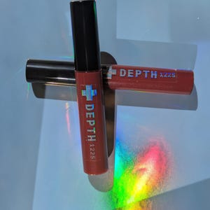May include: Two tubes of lip gloss with black caps and red tubes. The text "DEPTH 1225" is printed on the tubes. One tube is upright, and the other is lying horizontally. The background is a light blue surface with rainbow light reflections.