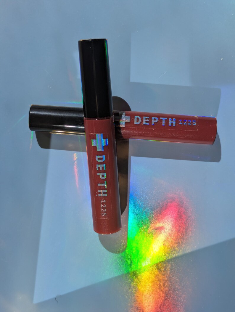 May include: Two tubes of lip gloss with black caps and red tubes. The text "DEPTH 1225" is printed on the tubes. One tube is upright, and the other is lying horizontally. The background is a light blue surface with rainbow light reflections.