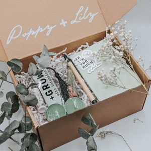 May include: A brown cardboard box with the words "Poppy + Lily" on the top. The box is open and contains a green journal, a pen, a small bottle, a sticker that says "If I'm Too Much", a bookmark that says "Grow with intention", and a few tealight candles.