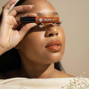 May include: A person holding a brown lipstick tube with the text "PAPI 1025" in front of their eye. The lipstick tube has a black cap and a white cross symbol. The person has brown skin, a white headband, and is wearing a beige top.