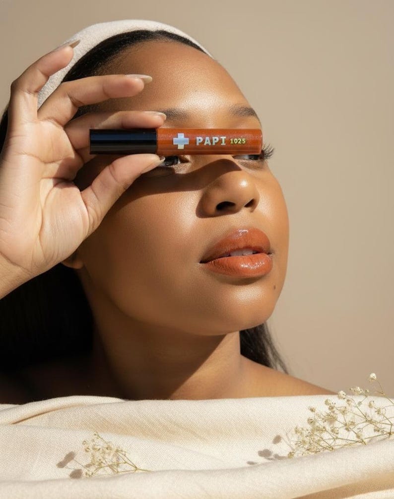 May include: A person holding a brown lipstick tube with the text "PAPI 1025" in front of their eye. The lipstick tube has a black cap and a white cross symbol. The person has brown skin, a white headband, and is wearing a beige top.