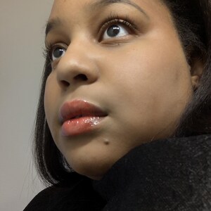May include: Close-up of a person's face, looking upwards. The skin has a warm tone, with glossy, light-colored lip gloss. The person has dark hair and is wearing a black top. The focus is on the eyes and lips.