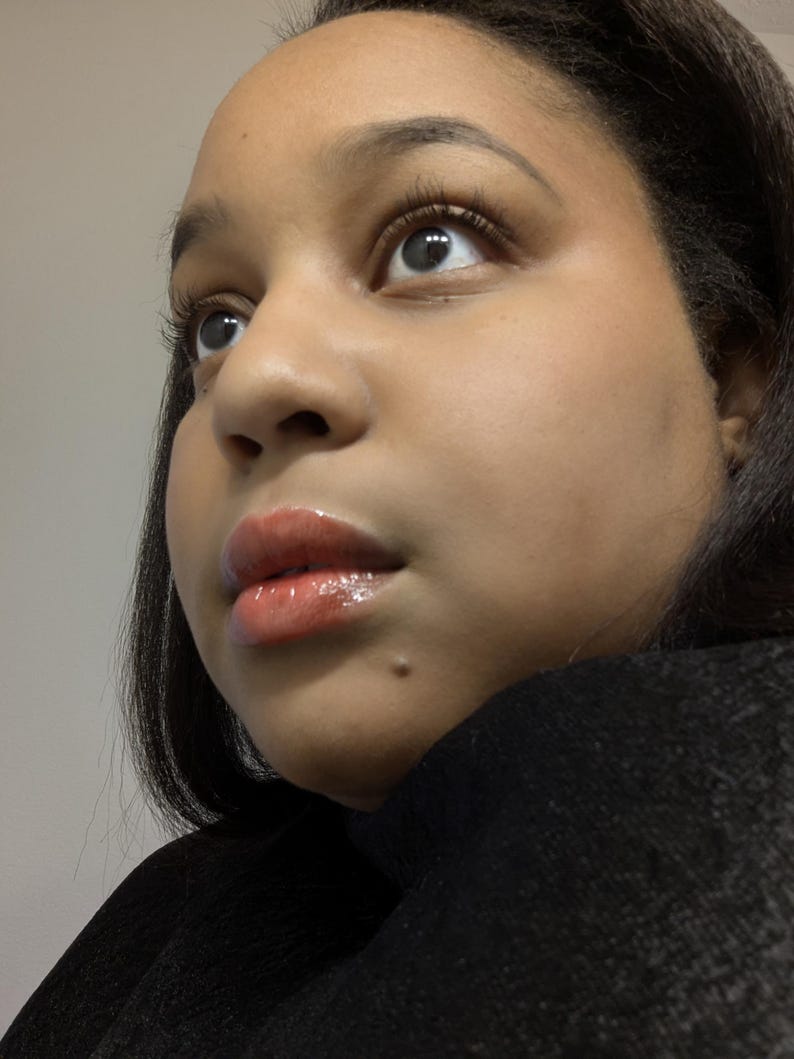 May include: Close-up of a person's face, looking upwards. The skin has a warm tone, with glossy, light-colored lip gloss. The person has dark hair and is wearing a black top. The focus is on the eyes and lips.