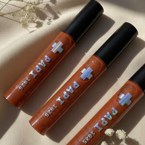 May include: Three tubes of lip gloss with a shimmering, reddish-brown hue. Each tube has a black cap and features the text "PAPI 1925" with a holographic cross symbol. The gloss tubes are arranged on a light-colored, textured surface.