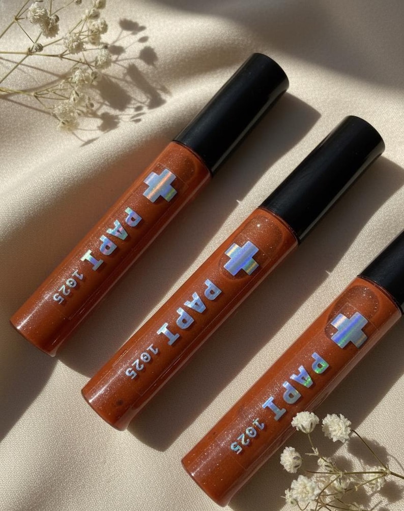 May include: Three tubes of lip gloss with a shimmering, reddish-brown hue. Each tube has a black cap and features the text "PAPI 1925" with a holographic cross symbol. The gloss tubes are arranged on a light-colored, textured surface.