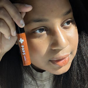 May include: Orange lip gloss tube with the text "PAPI 1625" and a white cross symbol. The tube has a black cap. The lip gloss is held up in front of a person's face.