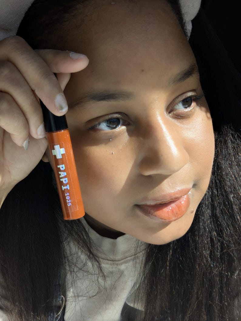 May include: Orange lip gloss tube with the text "PAPI 1625" and a white cross symbol. The tube has a black cap. The lip gloss is held up in front of a person's face.