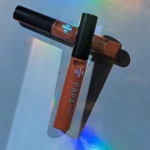 May include: Two tubes of lip gloss with black caps and orange-brown liquid. The tubes are labeled "PAPI 1925" in white text. The background has a rainbow-colored light effect.