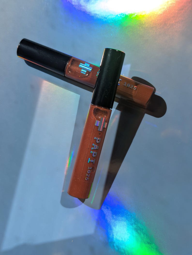 May include: Two tubes of lip gloss with black caps and orange-brown liquid. The tubes are labeled "PAPI 1925" in white text. The background has a rainbow-colored light effect.