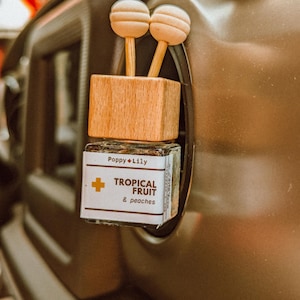 May include: A wooden car air freshener with a white and brown design. The air freshener has a label that says "Poppy + Lily Tropical Fruit & Peaches".