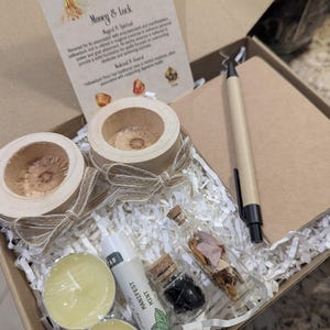 May include: A curated gift box with a spiritual theme. It contains two wooden candle holders, two small candles, a lip balm tube labeled "Manifest Mint", and two small glass bottles with cork stoppers. A pen and a card with text are also included.