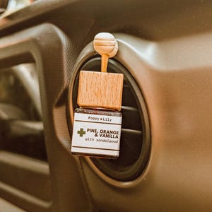 以下が含まれることがあります： 木製のキャップと白いプラスチッククリップが付いた小さな茶色のガラス瓶。瓶には「Poppy + Lily PINE, ORANGE & VANILLA with sandalwood」とラベルが貼られています。