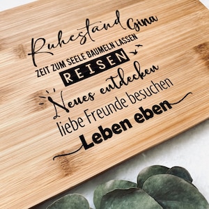 May include: A wooden cutting board with the text "Ruhestand Gina" and "Zeit zum Seele Baumeln Lassen" in black lettering. The text "Reisen" is in a larger font and is also in black. The text "Neues entdecken" and "liebe Freunde besuchen" is in a smaller font and is also in black. The text "Leben eben" is in a larger font and is also in black.