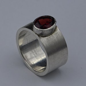 Könnte beinhalten: Ein silberner Ring mit breitem Band und einem oval geformten, tiefroten Edelstein in Zargenfassung. Der Ring hat eine gebürstete Metalloberfläche, die ihm ein strukturiertes Aussehen verleiht. Der Edelstein ist der Blickfang.