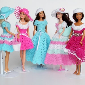 Op de afbeelding: Vijf gehaakte poppenjurken in roze, blauw en wit met bijpassende hoeden. De jurken zijn allemaal in verschillende stijlen en hebben ingewikkelde gehaakte details.