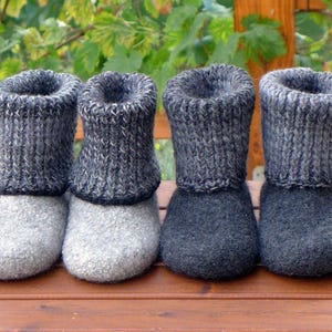 May include: Four pairs of gray and charcoal knitted wool slippers are arranged on a wooden surface. The slippers have a knitted cuff and a felted wool foot. The color palette is neutral, with shades of gray and charcoal.