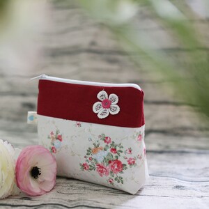 May include: A small red and white zippered pouch with a floral pattern and a white lace flower applique. The pouch is sitting on a wooden surface.