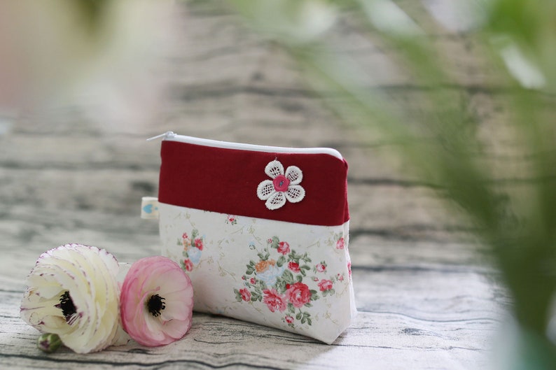 May include: A small red and white zippered pouch with a floral pattern and a white lace flower applique. The pouch is sitting on a wooden surface.