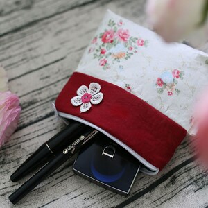 May include: A white and red floral fabric pouch with a zipper closure. The pouch is filled with makeup items, including a black eyeliner and a compact.