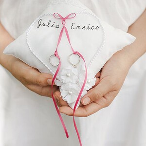 May include: A white heart-shaped pillow with the names "Julia" and "Enrico" embroidered on it. Two silver rings are placed on the pillow, tied together with a pink ribbon.