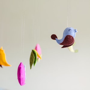 May include: A colorful felt mobile with a blue bird, green leaves, and a pink and yellow bird. The mobile is hanging from a white ceiling.
