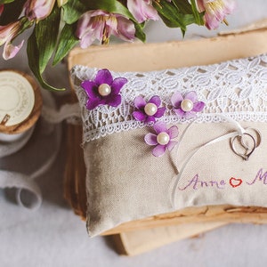 May include: A white pillow with lace trim and purple flowers with pearl centers. The pillow has the names "Anne" and "Mark" embroidered on it with a heart between the names. Two silver rings are on the pillow.