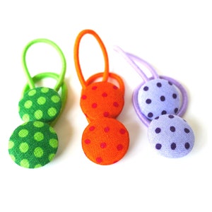 Puede incluir: Tres coleteros con botones de tela de lunares. Los coleteros son verde, naranja y morado.
