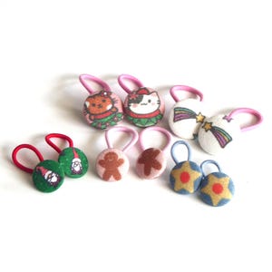 Puede incluir: Una colección de gomas para el pelo cubiertas de tela con varios diseños estampados, incluyendo gatos con suéteres, estrellas fugaces, hombres de jengibre, gnomos y estrellas. Las gomas tienen elásticos en tonos rosa, rojo y azul claro.