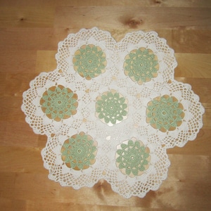 Puede incluir: Un tapete de ganchillo blanco hecho a mano con un diseño floral. El tapete presenta seis acentos florales verdes y un borde festoneado. El tapete está colocado sobre una superficie de madera.