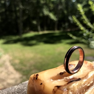 May include: A black and copper ring with a thin copper band and a wider black band. The ring is sitting on a piece of wood.