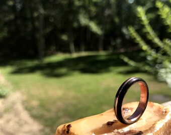 Puede incluir: Un anillo negro y cobre con una banda delgada de cobre y una banda negra más ancha. El anillo está sentado en un trozo de madera.