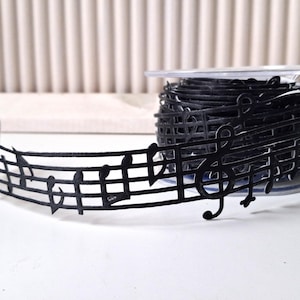 Könnte beinhalten: Eine Rolle schwarzes Notenband. Das Band ist in Form von Noten und Notenlinien geschnitten. Das Band befindet sich auf einer transparenten Kunststoffspule, wobei etwas Band im Vordergrund abgerollt ist.