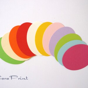 Könnte beinhalten: Eine Sammlung von elf glatten, matten Papier-Ovalen, die fächerförmig angeordnet sind. Die Farben umfassen Limettengrün, Creme, Orange, Rot, Off-White, Gelb, Blassrosa, Lavendel, Hellgrün, Himmelblau und Magenta. Der Text Cora Print ist sichtbar.