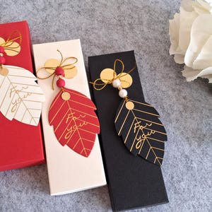 Könnte beinhalten: Rechteckige Geschenkboxen in Rot, Weiß und Schwarz, jede mit einem blattförmigen Anhänger. Die Anhänger tragen die Worte "For You" in goldener Schrift und sind mit goldenen Akzenten und Perlen verziert. Im Hintergrund eine weiße Blume.