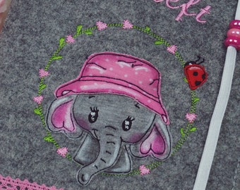 May include: Gray felt book cover with a pink and white embroidered elephant wearing a pink hat with white polka dots. The elephant is surrounded by a wreath of green leaves and pink flowers. The text "lil-Heft" is embroidered in pink on the cover.