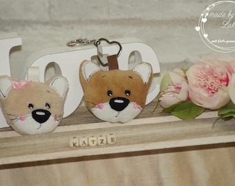 May include: Two plush cat keychains, one brown and one beige, with embroidered faces. The brown keychain has a heart-shaped ring and the beige keychain has a pink bow. The keychains are displayed on a wooden surface with the word "KATZE" spelled out in black tiles.