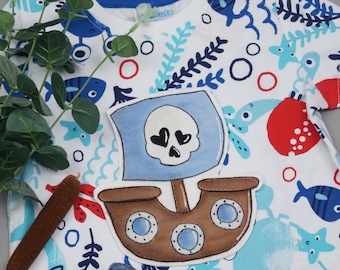 May include: A white t-shirt with a blue and brown pirate ship applique. The ship has a skull and crossbones on the sail and three portholes. The shirt has a blue, white, and red sea-themed pattern.
