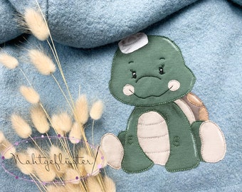 May include: A light blue fabric with a green turtle applique. The turtle has a white hat, a tan shell, and light tan accents. Dried bunny tail grass is in the foreground, and the text "Nahtgeflüster" is in a purple oval.
